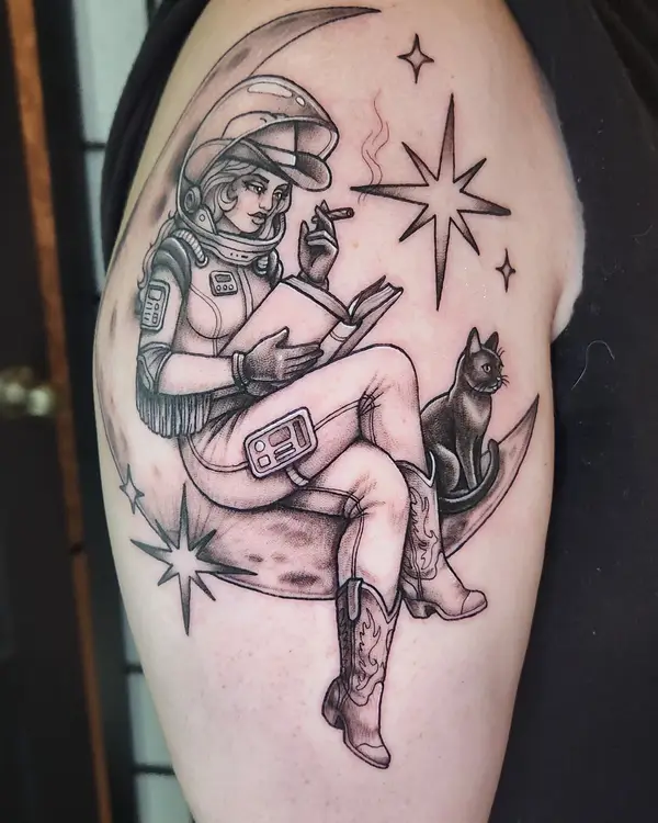 A black and grey tattoo of an astronaut in cowboy boots reading on a crescent moon beside a black cat