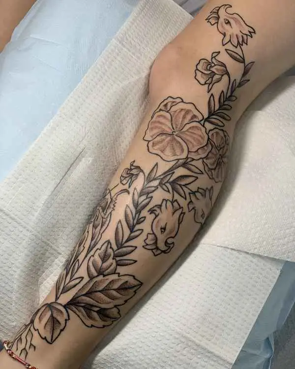 A black and grey illustrative leg piece of a small dragon winding through flowers and leaves, by Shannon Moxie