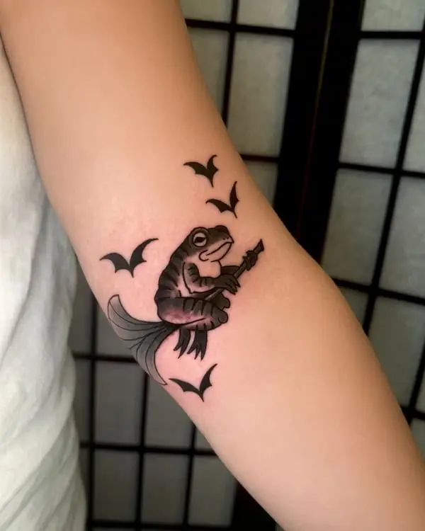 A black and grey tattoo of a toad riding a broomstick with bats, by Kate Elvey