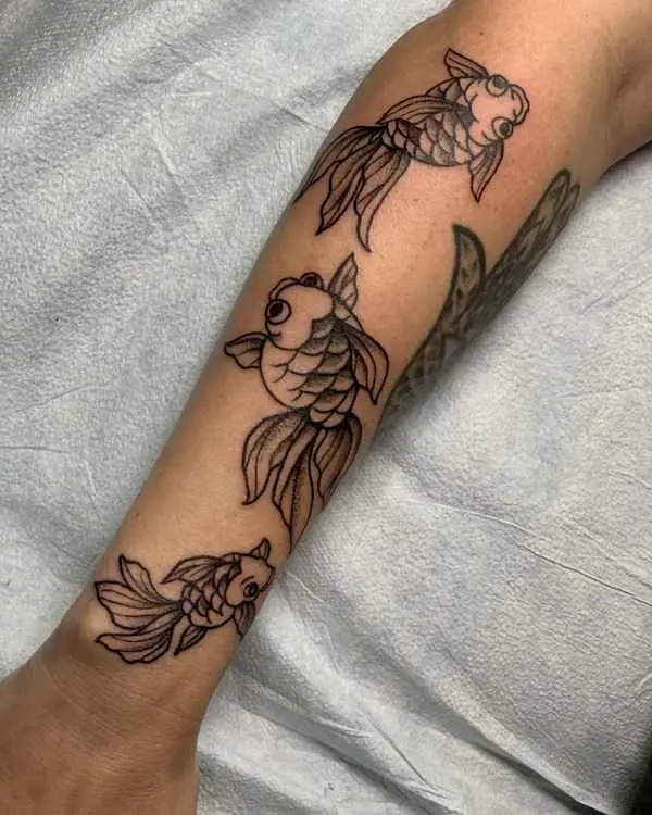 A black and grey tattoo of three goldfish swimming down a forearm, by Shannon Moxie