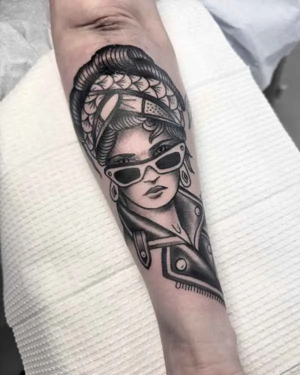 A black and grey tattoo of a woman in cat-eye sunglasses, a patterned headwrap, and a leather jacket, by Betty Rose