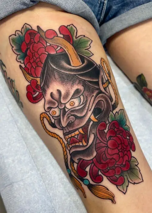 A neo-traditional Hannya mask tattoo with red flowers and a gold rope