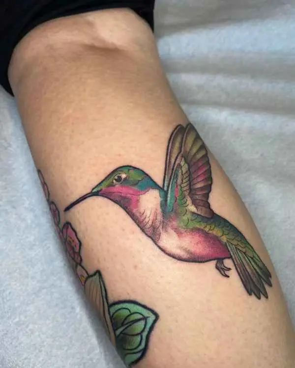A color tattoo of a hummingbird in flight with pink, green, and gold feathers, by Betty Rose